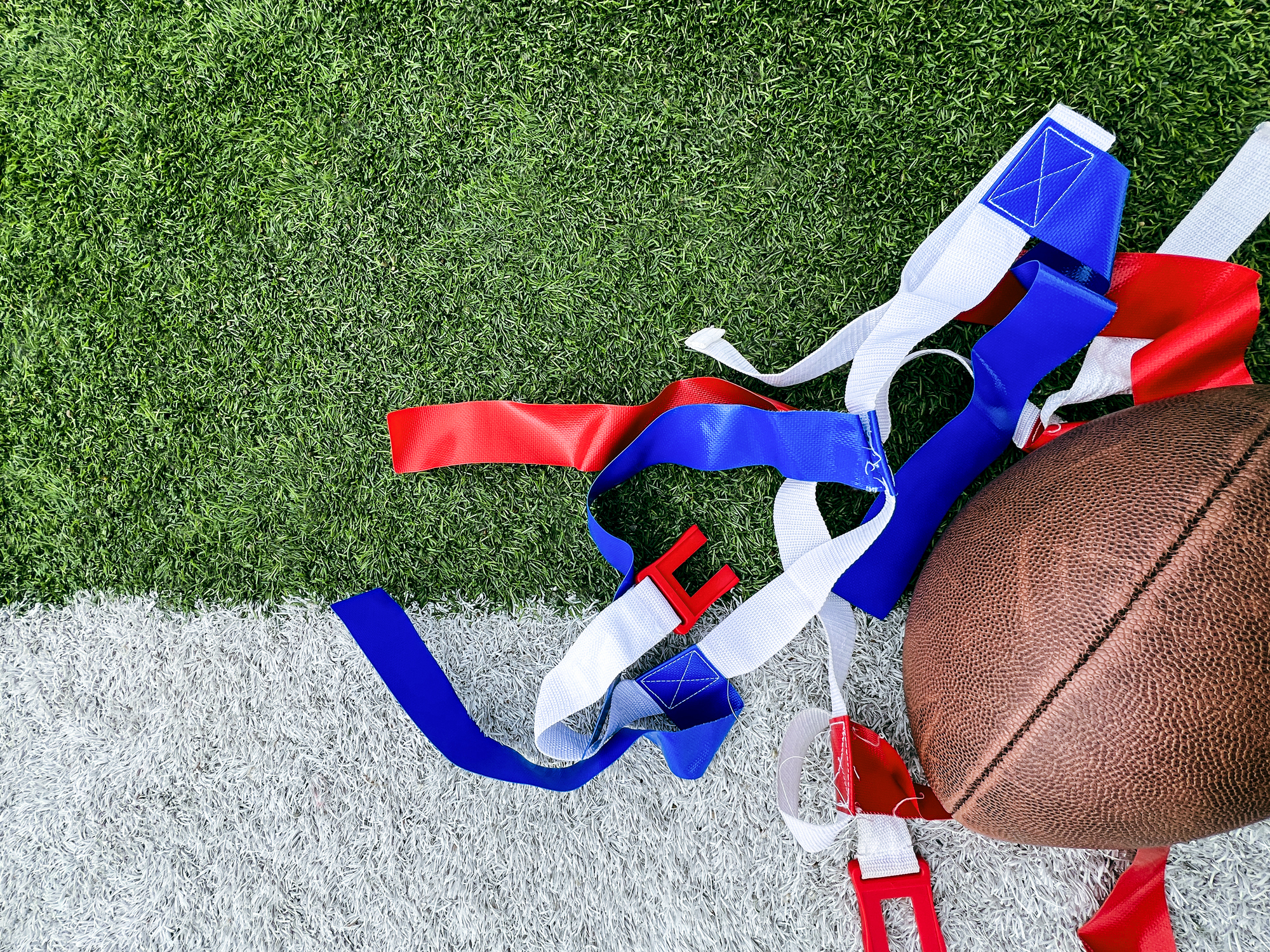 White belts with red and blue flags used for playing flag football. A football lays to the side. Set against the white and green turf of a football field.