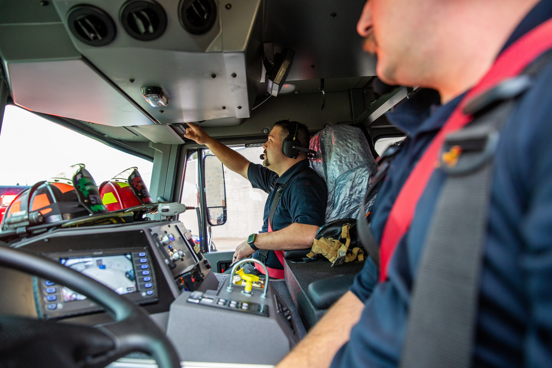 Two firefighters work inside the cab of a fire truck during an emergency response. A male firefighter in blue shirt and red suspenders operates the vehicle controls from the driver's position, while a female firefighter in dark uniform sits nearby managing communication and monitoring equipment. The cab interior shows multiple control panels, gauges, and emergency equipment mounted on the dashboard and overhead. Emergency gear and equipment are visible on the truck exterior through the windows. The scene captures real-time operational procedures during active emergency dispatch.