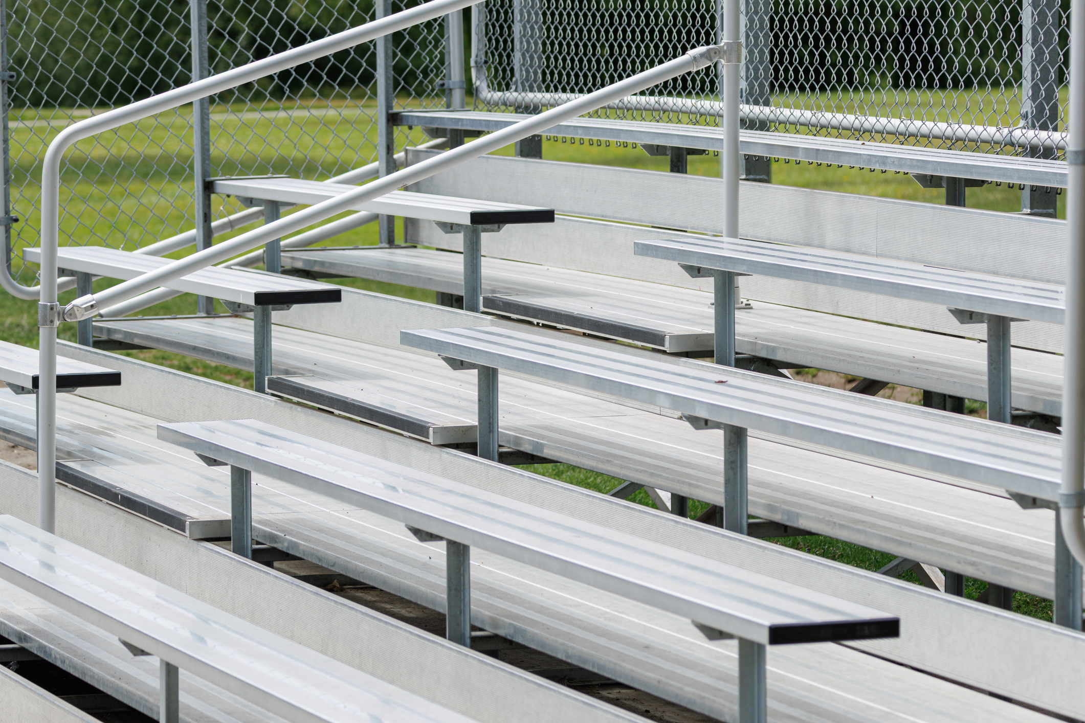 Empty aluminum bleachers arranged in parallel rows at an outdoor sports field, showing metal seating, handrails and a chain link fence with green grass beyond, evoking quiet anticipation before a game.